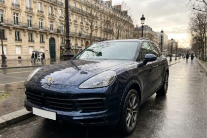 Porsche Cayenne bleu nuit garée sur un boulevard parisien sous la pluie, reflet sur le bitume mouillé, photographie éditoriale réaliste pour un article sur l'assurance sur mesure